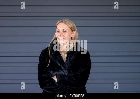Eine blonde Frau in einem kuscheligen schwarzen Pelzmantel steht mit überkreuzten Armen. Ihr Ausdruck wird durch ein dezentes Lächeln gemildert. Stockfoto