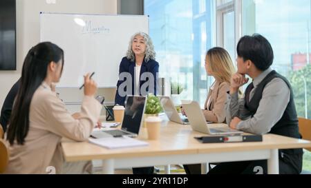 Eine leitende asiatische CEO leitet ein Treffen mit ihrem Team, bei dem Strategien für eine neue Marketingkampagne diskutiert werden. Verschiedene asiatische Geschäftsleute sitzen A Stockfoto