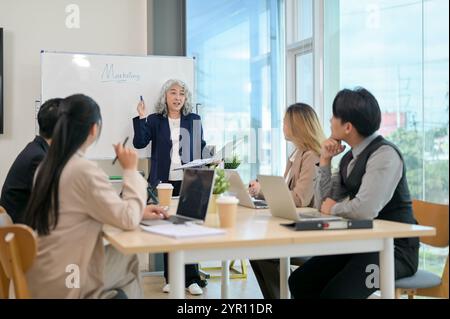 Eine leitende asiatische CEO leitet ein Treffen mit ihrem Team, bei dem Strategien für eine neue Marketingkampagne diskutiert werden. Verschiedene asiatische Geschäftsleute sitzen A Stockfoto