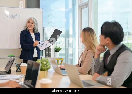 Eine leitende asiatische CEO leitet ein Treffen mit ihrem Team, bei dem Strategien für eine neue Marketingkampagne diskutiert werden. Verschiedene asiatische Geschäftsleute sitzen A Stockfoto