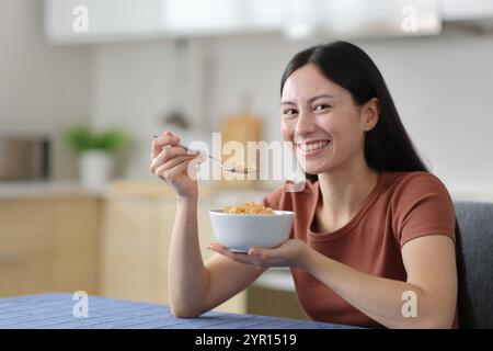 Glückliche asiatische Frau isst Müsli und sieht dich in der Küche zu Hause an Stockfoto