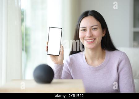 Glückliche asiatische Frau mit virtuellem Lautsprecher, der zu Hause einen leeren Telefonbildschirm zeigt Stockfoto