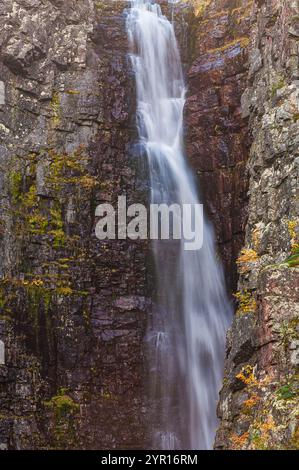 Ein atemberaubender Wasserfall fließt anmutig die steilen Klippen aus zerklüftetem Stein hinunter. Leuchtende Herbstblätter bilden einen farbenfrohen Kontrast zum stimmungsvollen Felsen Stockfoto