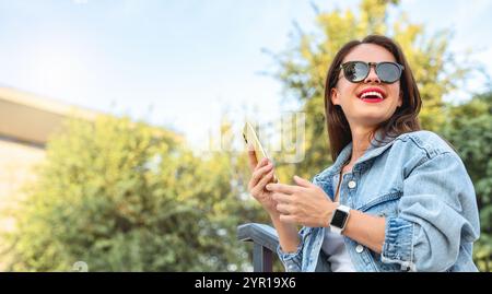 Junge kaukasische Frau in Jeansjacke und Sonnenbrille, die ihr Smartphone hält und draußen lächelt. Stockfoto