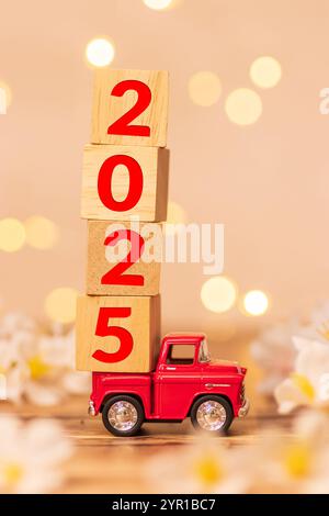 Rotes Truck-Spielzeug mit Holzwürfel mit der Nummer 2025 auf der Rückseite des Pickups für den Beginn des neuen Jahres 2025 mit unscharfem weißem Blumenfeld Stockfoto