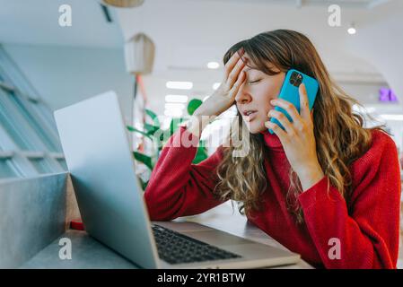 Eine gestresste Geschäftsfrau, die telefoniert und einen Laptop benutzt, während sie im Coworking Space arbeitet, die Stirn mit der Hand berührt und schlechte Nachrichten hat Stockfoto