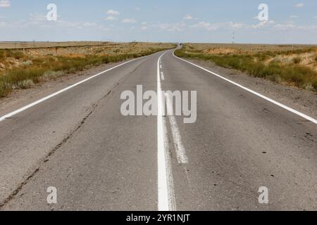 Eine lange, gerade Autobahn führt durch das weite, offene Gelände Kasachstans, umgeben von trockenem Gras und blauem Himmel an einem sonnigen Tag. Stockfoto