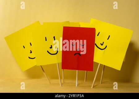 Rotes Papier mit traurigem deprimiertem Gesicht, umgeben von gelben lächelnden Papieren, Konzept der Depression und Isolation von der Gesellschaft Stockfoto