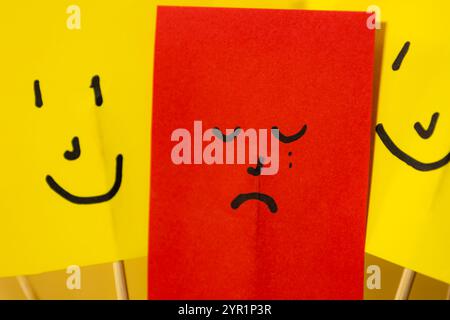 Nahaufnahme von rotem Papier mit traurigem depressivem Gesicht, umgeben von gelben Smiley-Papieren, Konzept von Depression und Isolation von der Gesellschaft Stockfoto