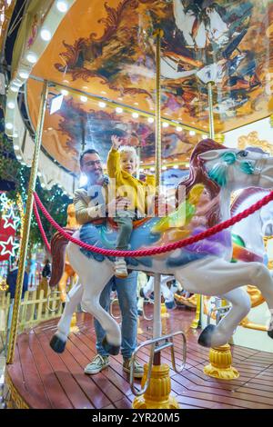 Vater und Sohn genießen eine Karussellfahrt auf einem weihnachtsmarkt, winken und lächeln, was eine fröhliche Erinnerung an die Feiertage schafft Stockfoto