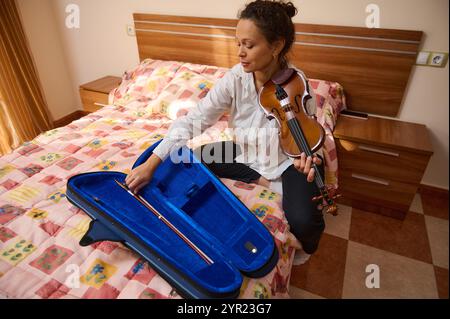 Eine Frau sitzt auf einem Bett in einem gemütlichen Schlafzimmer und bereitet ihre Geige vor. Sie legt den Bogen vorsichtig in die Hülle und reflektiert einen Moment persönlicher Kreativität und Stockfoto
