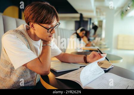 Universitätsstudent mit Brille studiert fleißig in einer stilvollen Bibliotheksumgebung, umgeben von Büchern und Papieren, reflektiert Konzentration und Stockfoto
