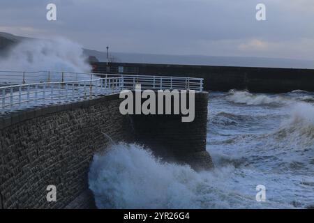 Aberystwyth Wales UK Wetter 2. Dezember 2024. Ein kalter, stürmischer Wintermorgen an der walisischen Küste, große Wellen krachen über die Seeschützung und Hafenlicht mit rosa Himmel über der Küste, Credit: mike davies/Alamy Live News Stockfoto