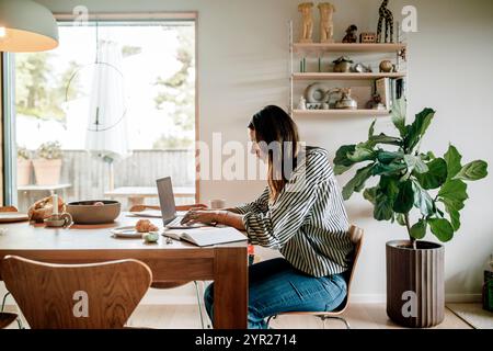 Seitenansicht einer fokussierten Geschäftsfrau, die am Laptop arbeitet, während sie zu Hause neben dem Esstisch sitzt Stockfoto
