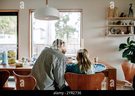 Rückansicht Mutter, die den Sohn bei den Hausaufgaben begleitet, während er zu Hause auf einem Stuhl neben dem Tisch sitzt Stockfoto
