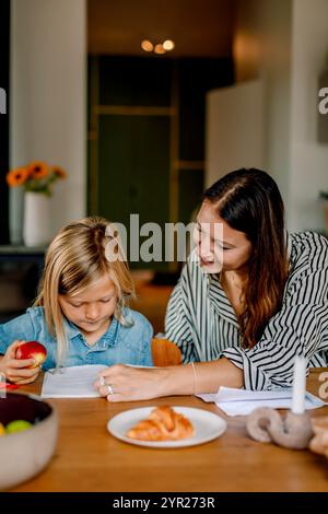 Mutter, die den Sohn bei den Hausaufgaben begleitet, während sie zu Hause neben dem Tisch sitzt Stockfoto