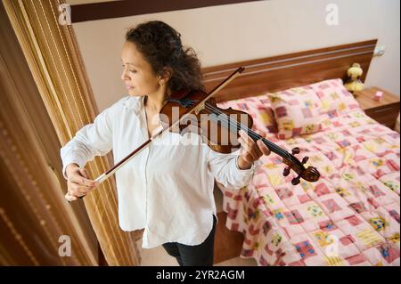 Eine junge Frau übt das Geigenspiel in einem warmen, gemütlichen Schlafzimmer. In einem weißen Hemd konzentriert sie sich auf ihre Musik, mit einem gemusterten Bett und weich Stockfoto