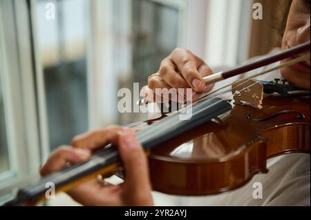 Eine Nahaufnahme eines Musikers, der in der Nähe eines sonnendurchfluteten Fensters Geige spielt, betont die beredte Bewegung des Bogens über die Streicher und das komplizierte d Stockfoto