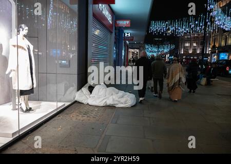 Fußgänger passieren einen Obdachlosen, der in einer Bettdecke vor einem Schaufenster in der Oxford Street im Zentrum von London schläft Stockfoto