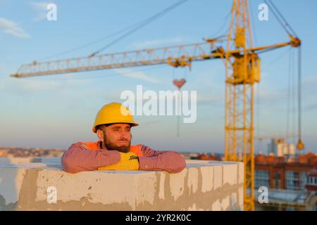 Bauarbeiter mit Sicherheitsweste und Schutzhelm, der sich an sonnigen Tagen mit Kran im Hintergrund an der Wand lehnt Stockfoto