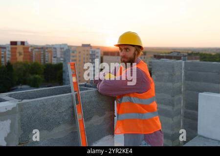 Porträt eines selbstbewussten Bauarbeiters mit Schutzhelm und Sicherheitsweste, der bei Sonnenuntergang auf Betonblock geneigt ist Stockfoto