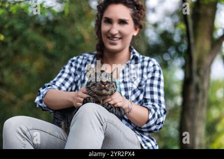 Die junge Frau sitzt friedlich in einem üppigen Wald, hält eine Tabbykatze auf ihren Beinen und strahlt Liebe und Sorgfalt aus Stockfoto