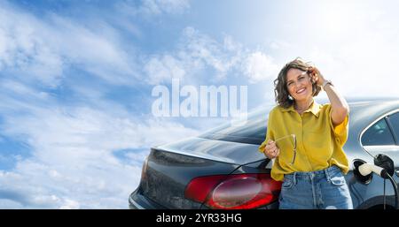 Frau mit gebogenen Haaren, die ein gelbes Hemd trägt, steht neben dem Aufladen des Elektroautos. Windräder im Hintergrund. Stockfoto