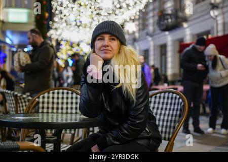 Eine Winterszene mit einer Frau, die eine Tasse heißes Getränk in einem gemütlichen Straßencafé genießt. Der Hintergrund zeigt eine festliche Straße, die von New beleuchtet wird Stockfoto