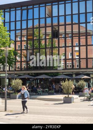 Ein hohes Spiegelglasgebäude spiegelt ein Backsteingebäude in seinem Glas wider, in dem die Leute in einem Straßencafé sitzen und den Sommertag genießen. Stockfoto