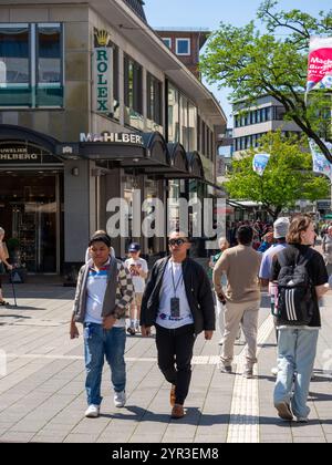 An einem sonnigen Sommertag laufen Menschenmassen die Einkaufsstraße Holstenstraße hinunter mit der Beschilderung eines Rolex-Geschäfts im Hintergrund. Stockfoto