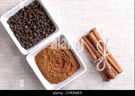Cassia Zimtpulver und Knospen in weißen Schüsseln, mit Zimtstäbchen zu einem Bündel zusammengebunden, auf Leinengewebe und mit leerem Platz für Text. Stockfoto