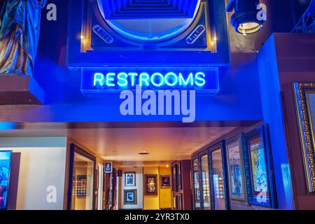 Neonblaue Toiletten-Schild, das im schwach beleuchteten Flur des Hard Rock Cafe leuchtet, mit gerahmten Plakaten auf dem Hintergrund der Wände. New York. USA. Stockfoto