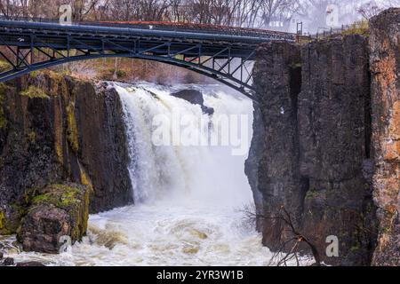 Der Wasserfall Great Falls in Paterson, New Jersey, stürzt mächtig über felsige Klippen mit einer eisernen Brücke darüber. Stockfoto