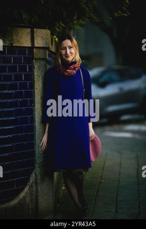 Eine Frau in einem Mantel und Halstuch steht an einer Wand, hält ihre Baskenmütze in der Hand, auf einer Straße im Herbst. Stockfoto