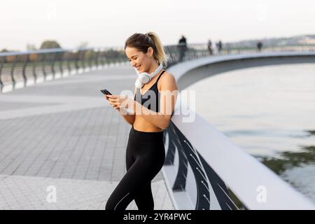 Eine Joggerin hält an ihrem Telefon am Fluss an, trägt Trainingsausrüstung und Kopfhörer und genießt die warme Sonne und die ruhige Atmosphäre Stockfoto