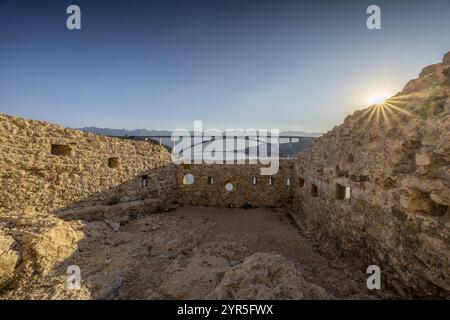 Blick auf die steinige Ruine mit der Brücke Pag im Hintergrund, Sonnenaufgang über den Bergen, Pag, Zadar, Kroatien, Europa Stockfoto