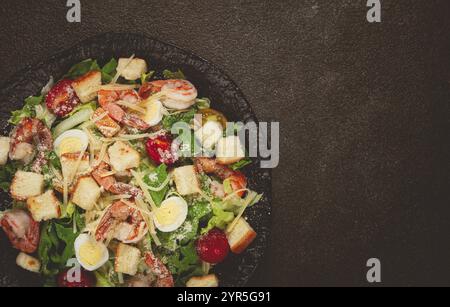 Caesar-Salat mit Tigergarnelen, Garnelensalat, Salatblättern, Wachteleiern, Kirschtomaten, parmesankäse, auf einem schwarzen Teller Stockfoto