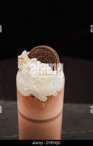 Schokoladenmilchshake mit Schlagsahne, serviert in einem Glas auf einem dunklen Tisch. Schokoladenkekse drauf. Dreifachgetränk. Stockfoto