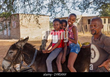 Afrika-Dorf, afrikanische Kinder, die auf dem Bauernhof auf einem Esel reiten, Vater, der das Vieh hält, ländliche Häuser im Hintergrund Stockfoto