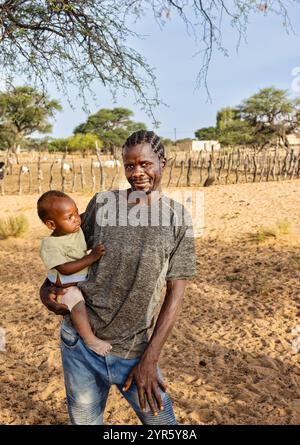 Ein afrikanischer Dorfmann hält ein Baby auf der Farm vor dem Viehstall Stockfoto