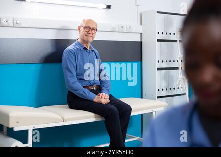 Pensionierter Mann bequem auf Krankenhausbett sitzt wartet auf die Untersuchung in der Gesundheitsklinik. Portait eines älteren Patienten in Arztpraxis bereit für den Arztbesuch. Stockfoto