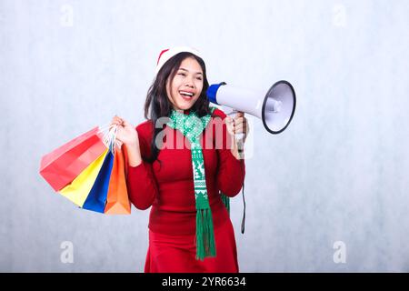 Charmante junge indonesien-Frau mit rotem weihnachtspullover, weihnachtsmütze und Schal, glücklicher freimütiger Hand, die Megaphon-Lautsprecher hält und Farbe trägt Stockfoto