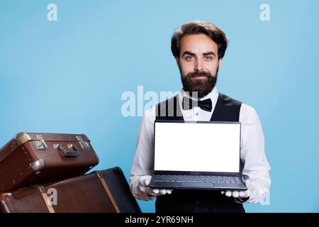 Der schicke Portier zeigt im Studio einen weißen Bildschirm auf dem Laptop, während er weiße Handschuhe neben einem Stapel von Trolley-Taschen trägt. Professionelle Pagen, die in einem luxuriösen Hotel arbeiten, präsentieren leere Displays. Stockfoto