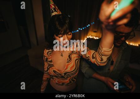 Eine junge Frau mit gemustertem Oberteil und Partyhut tanzt fröhlich bei einer lebhaften Geburtstagsfeier, umgeben von festlichen Lichtern und Ambiente. Stockfoto