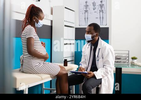 Schwangere schwarze Frau sitzt auf dem Krankenhausbett und spricht mit dem Arzt über ihre Mutterschaftsuntersuchung, während sie Schutzmasken trägt. Die werdende Mutter hört aufmerksam auf den afroamerikanischen Arzt. Stockfoto