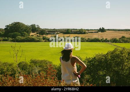 Junge Frau mit weißem Hut, die an einem sonnigen Sommertag ein schönes grünes Feld mit einem Bauernhof im Hintergrund in santarem, portugal, bewundert Stockfoto