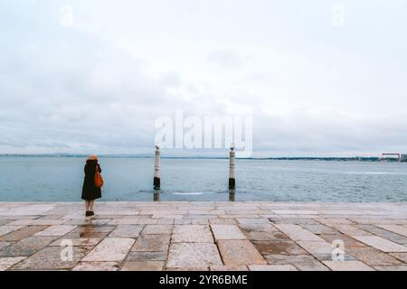 Junge Frau, die an einem bewölkten Tag auf dem Pier von cais das colunas in lissabon steht und den tejo und die zwei Säulen, die aus dem Wasser auftauchen, bewundert Stockfoto