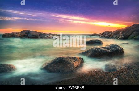 Sanfte Wellen, die bei Sonnenuntergang über Felsen am Strand strömen, erzeugen einen wunderschönen Bewegungsunschärfeeffekt mit leuchtenden Farben am Himmel Stockfoto