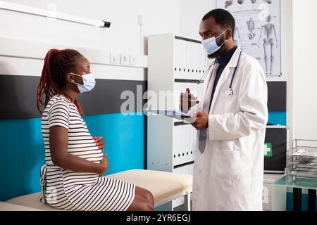 Schwangere Dame mit Gesichtsmaske, die auf den professionellen männlichen Arzt im Arztzimmer hört. afroamerikanischer Arzt, der schwarze Frau, die auf Krankenhausbett sitzt, Mutterschaftsratschläge gibt. Stockfoto