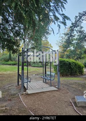 Metallschaukel unter einer Pergola in einer ruhigen Parklandschaft, umgeben von Grün und Bäumen. Ein ruhiger Ort für Entspannung und Outdoor-Genuss. Stockfoto